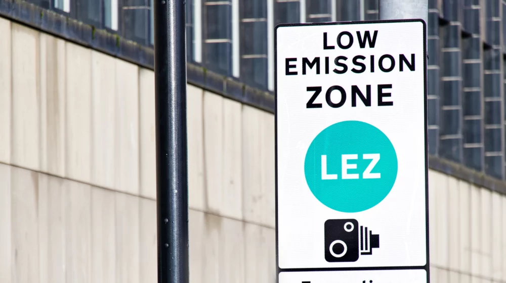 A sign indicating a Low Emission Zone (LEZ) in an urban area. The sign features a bold, turquoise circle with the letters ‘LEZ’ in white and an icon of a camera with a flash below the text. The background shows a blurred modern building facade.