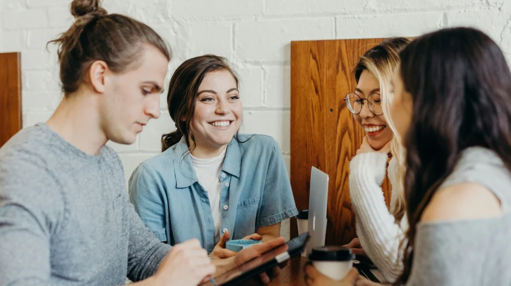 Three young adults sitting at a table with laptops, notebooks and drinks and laughing.