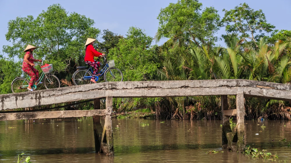 Two Vietnamese women wearing traditional conical hats and vibrant floral pants are riding bicycles across a narrow concrete bridge over a river. The background features lush greenery and palm trees under a clear blue sky.