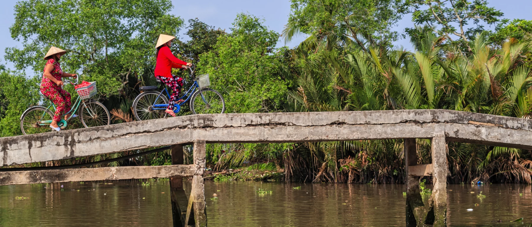 Two Vietnamese women wearing traditional conical hats and vibrant floral pants are riding bicycles across a narrow concrete bridge over a river. The background features lush greenery and palm trees under a clear blue sky.
