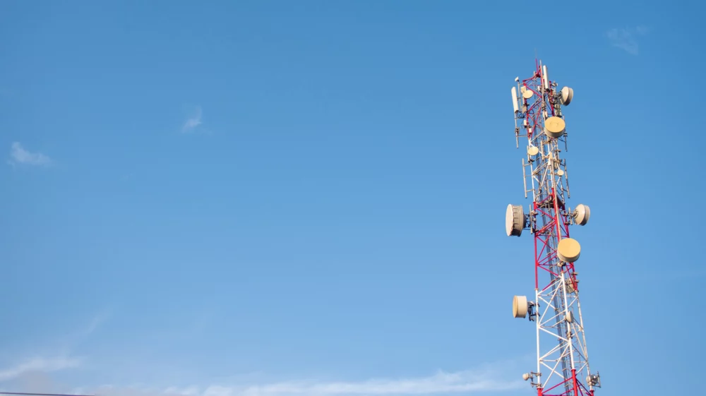 A wide-angle view of a tall communications tower equipped with various satellite dishes and antennas, set against a clear blue sky with minimal cloud cover.