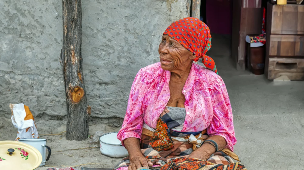 African older woman sitting on the floor and eating her lunch in front of a village house.