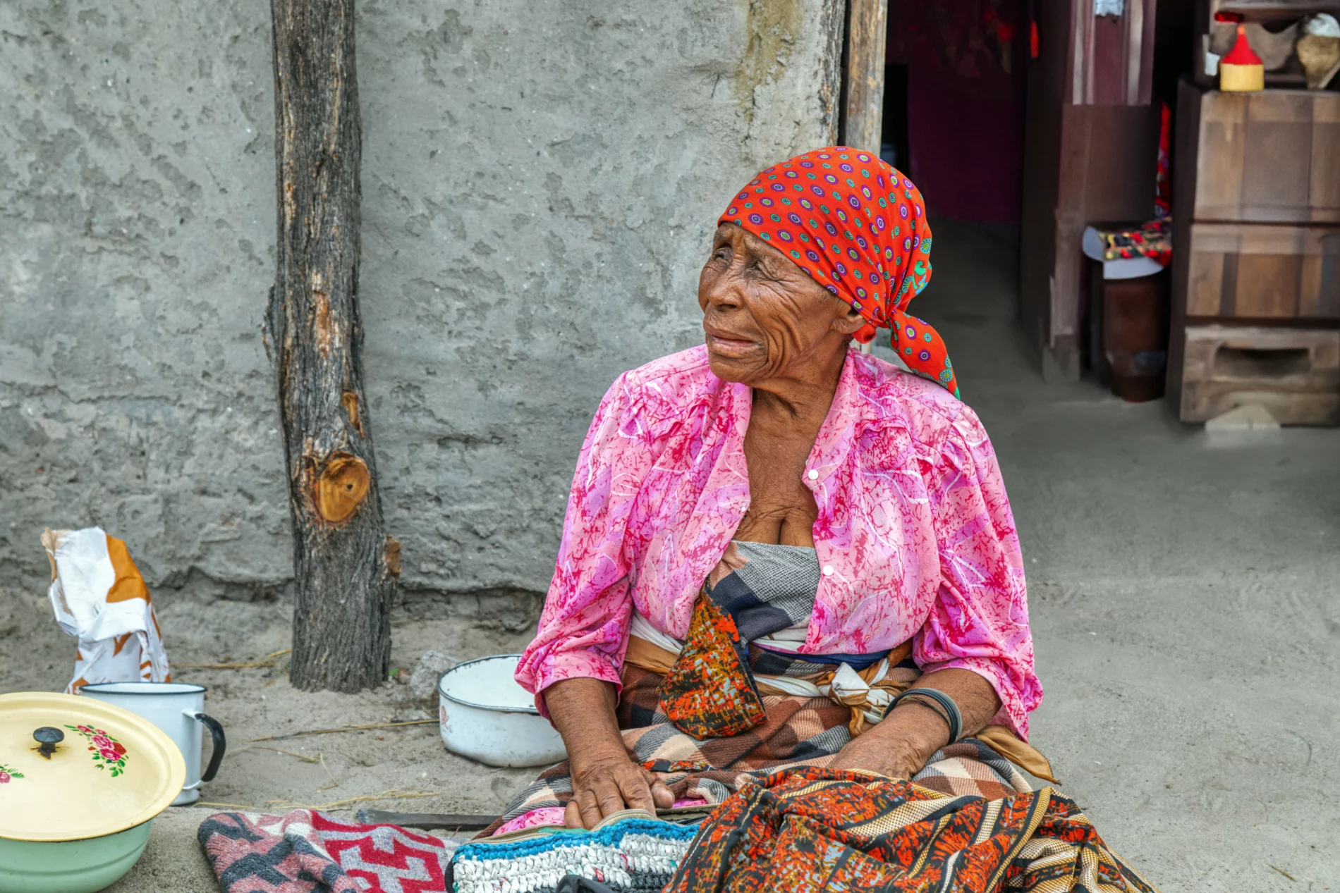 African older woman sitting on the floor and eating her lunch in front of a village house.
