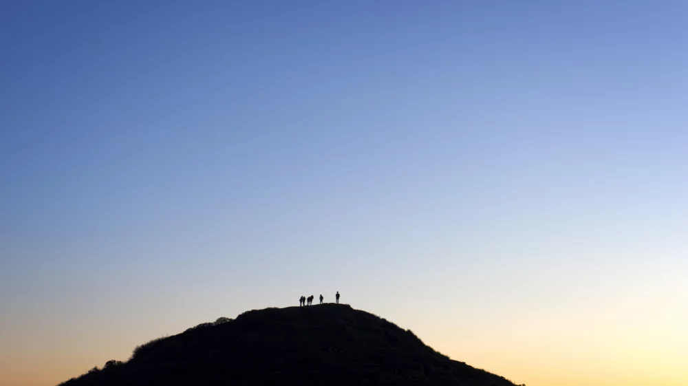 A dark blue sky above the backlit silhouette of a hill with several people standing at the peak, very tiny on top of the hill.