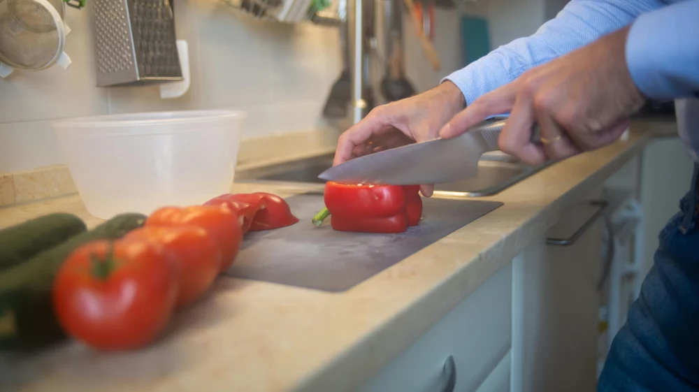 Two hands slicing a paprika on a thin, gray, plastic chopping board on a kitchen counter in front of a sin. Three tomatoes and two cucumbers are visible on the counter.