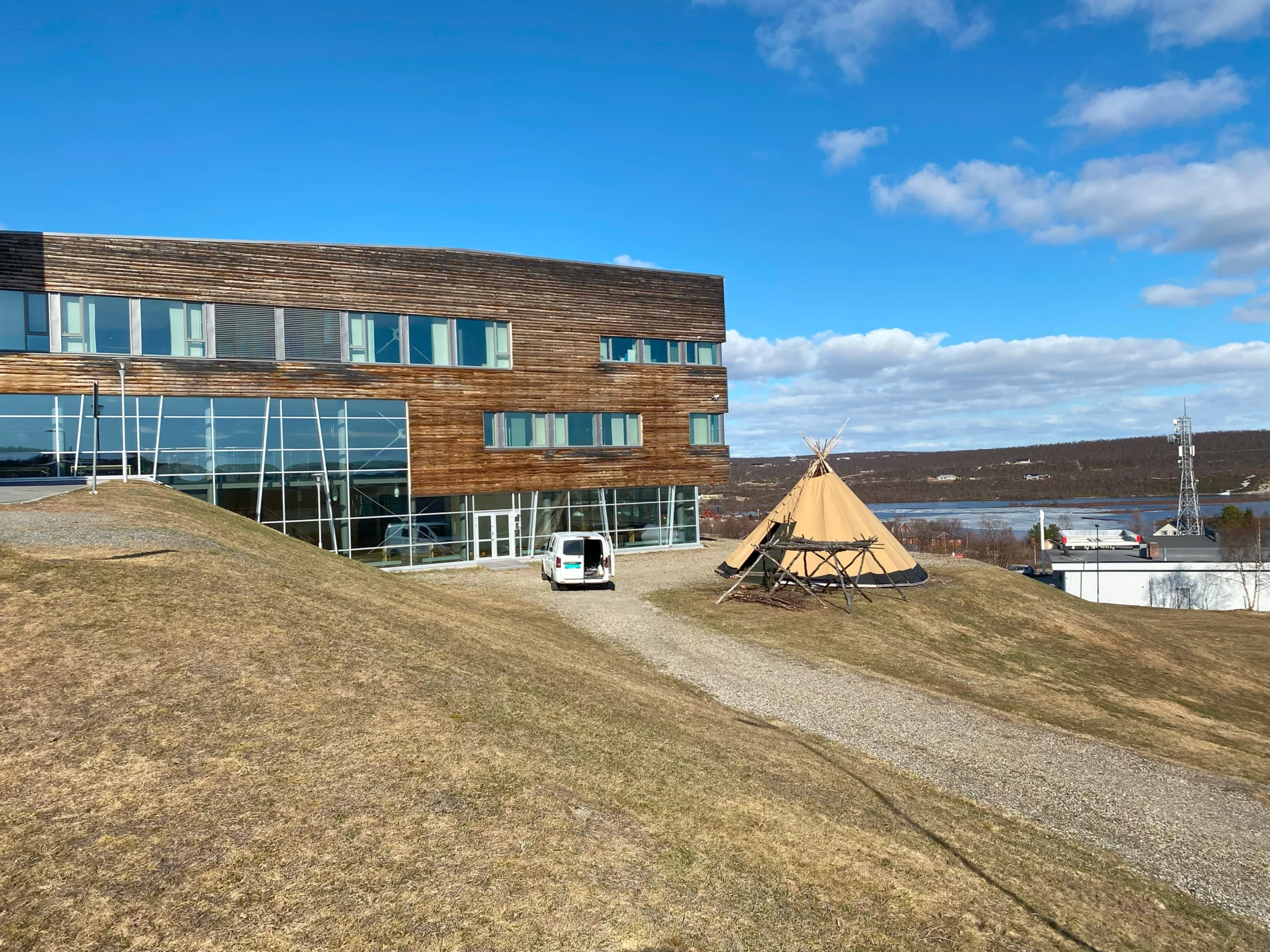 Second half of the modern wood and glass university building as seen from a hill on which the entrance floor sits. A small van is parked in front as well as a flat cone shaped tent. A body of water and a wooded hill can be seen in the background.