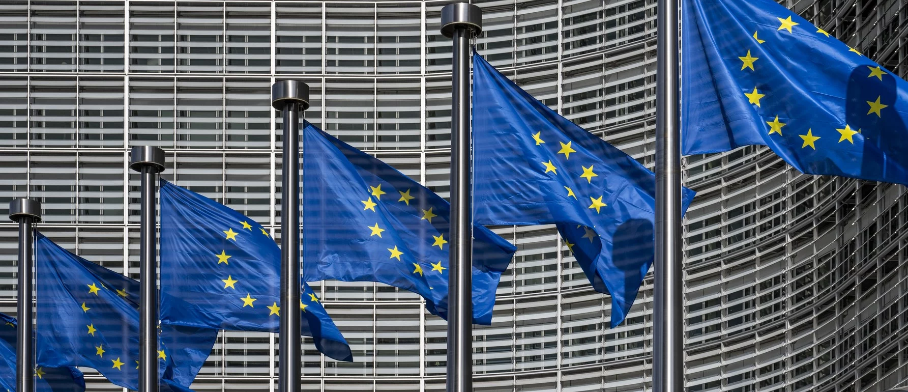 EU flags are mounted on tall flagpoles and are fluttering in the wind in front of the European Comission's large, modern headquarters. The building’s facade is composed of numerous horizontal and vertical lines, creating a grid-like pattern in the background.