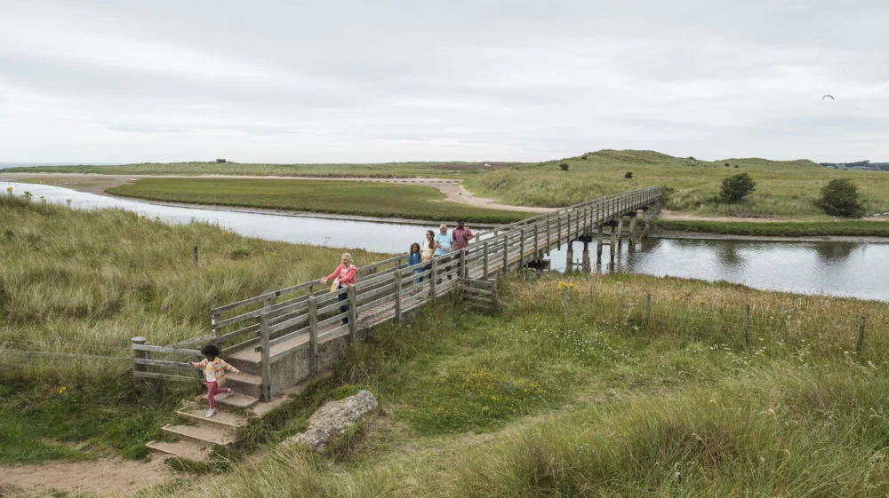 A group of people, including children, walk across a wooden footbridge over a river in a grassy landscape under an overcast sky.