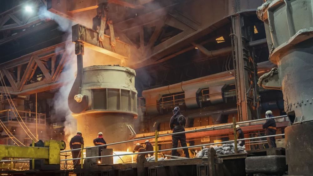 A huge metal vat, suspended from enormous hooks glows with the molten steel it contains. Below, people in protective gear stand ready for the steel to pour into a mould.