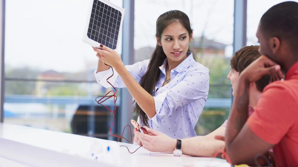 A young South Asian woman stands at a desk holding up a medium-sized photovoltaic cell, showing it to two other people who have their backs to the camera.