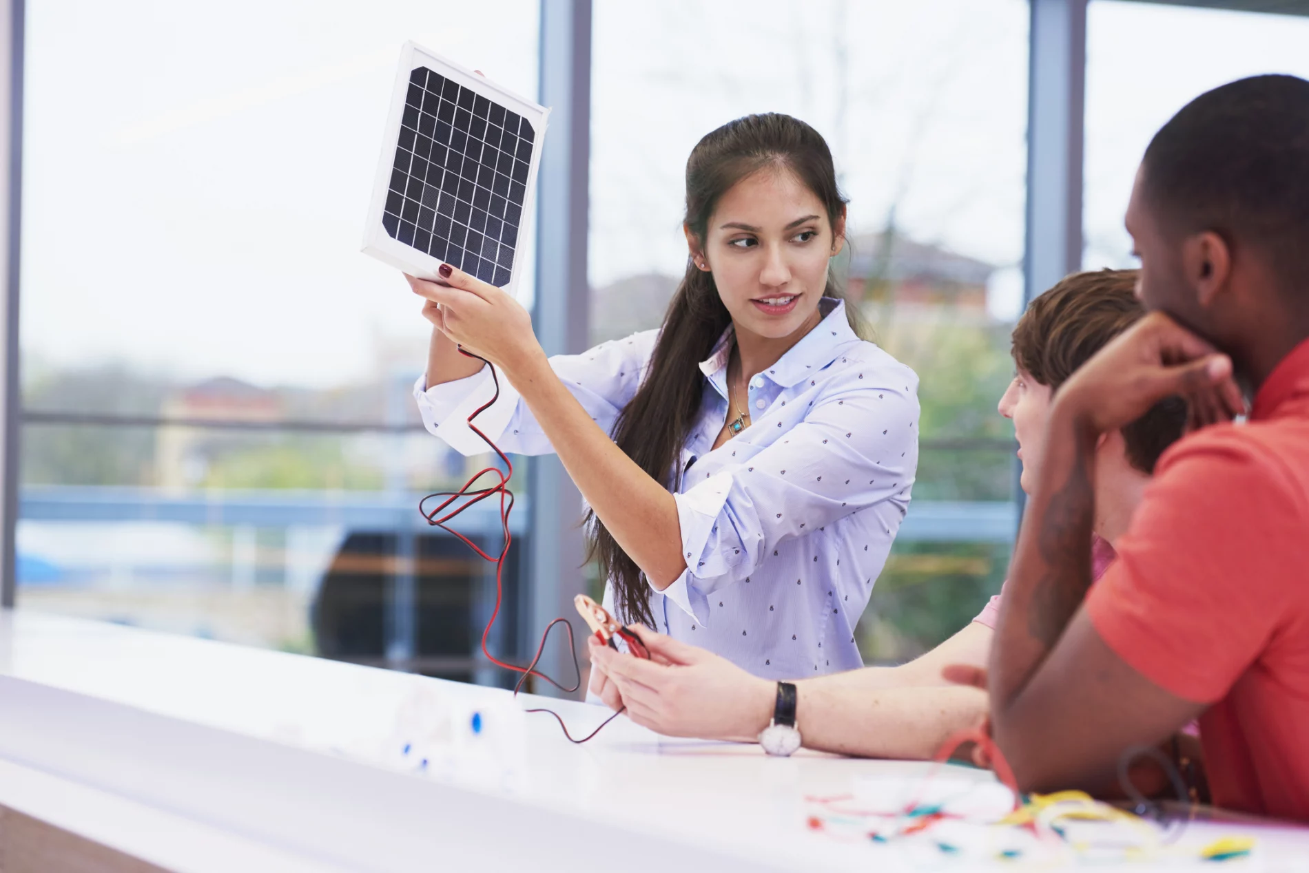 A young South Asian woman stands at a desk holding up a medium-sized photovoltaic cell, showing it to two other people who have their backs to the camera.
