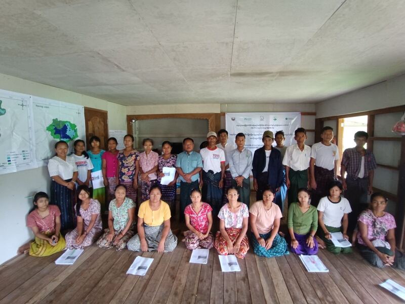 A group photo of community members participating in a biodiversity conservation training in Maing Naung village. The group includes both men and women of various ages, with some standing in the back and others sitting on the wooden floor in the front row. They are holding documents related to the training.