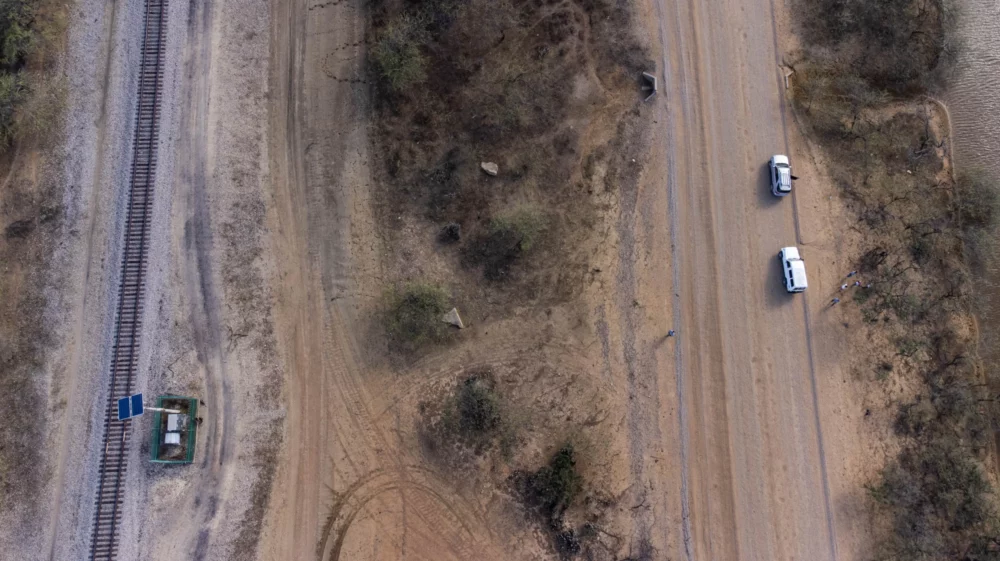 Aerial view of a barren landscape in La Guajira, featuring two white vehicles driving on a dirt road parallel to a railway track. A small solar panel installation is visible near the railway.