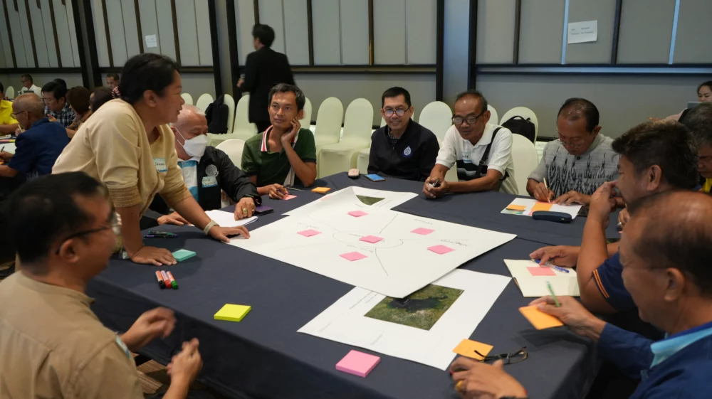 A participant stands and shares her thoughts with a group during a discussion session. Several people sit around a table covered with papers, maps, and sticky notes, listening attentively.