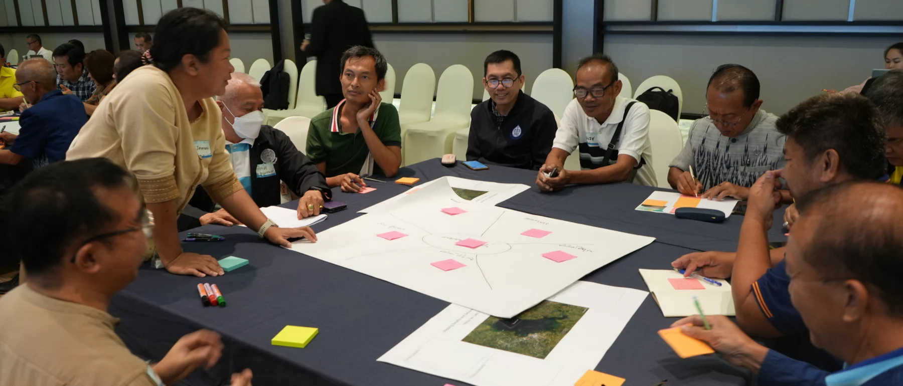 A participant stands and shares her thoughts with a group during a discussion session. Several people sit around a table covered with papers, maps, and sticky notes, listening attentively.
