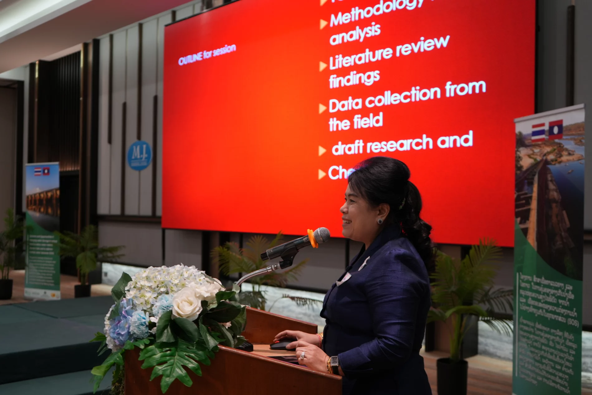 Dr. Ninphaseuth Xayaphonesy, Director General of the Department of Women in Development at the Lao Women’s Union, presents a slide on gender analysis and the Sustainable Development Goals (SDGs) with a focus on Gender Equality, Disability, and Social Inclusion (GEDSI). She stands at a podium with a microphone, speaking to an audience, with a large red presentation slide displayed in the background.