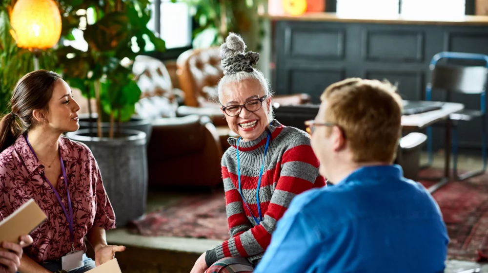 A stylishly dressed older woman laughs, as she sits in a group with other people.