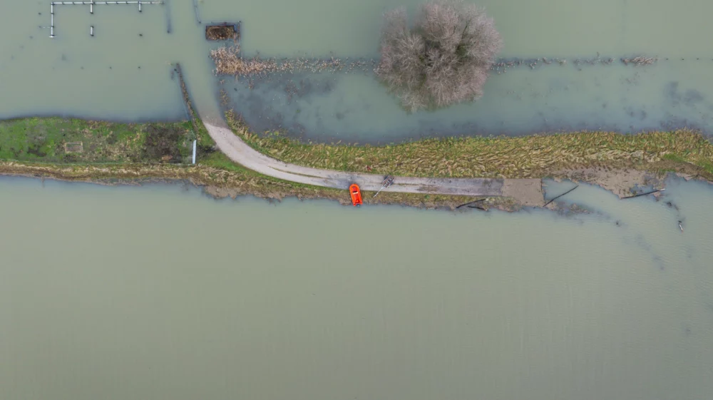 Aerial view of a narrow road cutting through a flooded landscape, with a single orange vehicle parked on the road near a large, leafless tree surrounded by water.