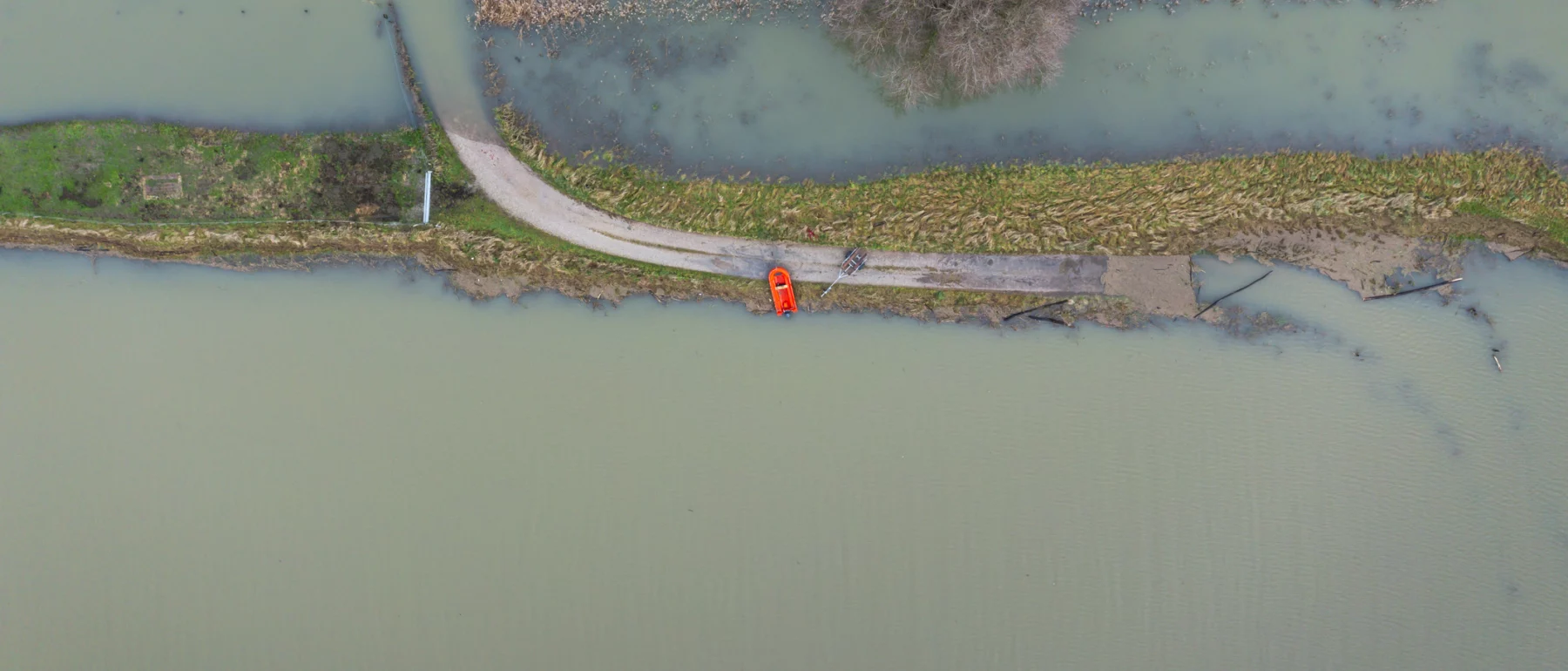 Aerial view of a narrow road cutting through a flooded landscape, with a single orange vehicle parked on the road near a large, leafless tree surrounded by water.