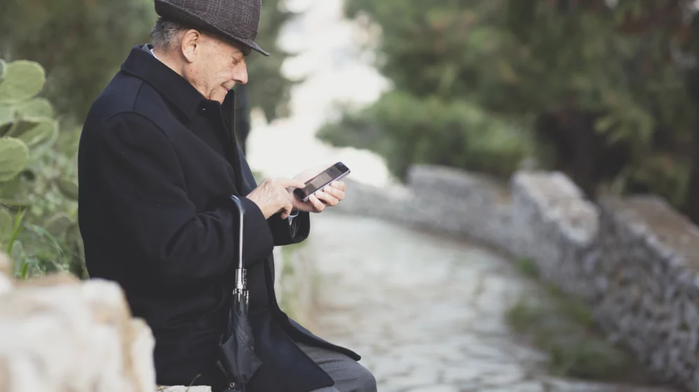 Portrait of an older man. He is sitting on a wall on the edge of a stone path. He is wearing a dark tweed hat and a black wool coat. He is using his right index finger to use his smartphone which he holds in his left hand. A black umbrella is hooked over his right wrist. Soft focus greenery makes up the background.