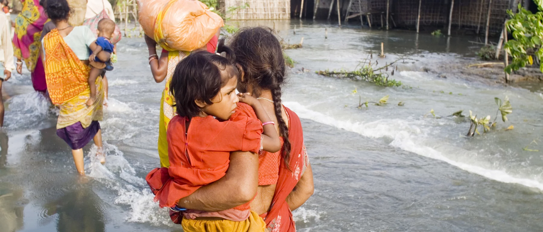 A woman in an orange sari carries a child on her left hip through flood waters, Kosi River flood of Bihar, 2008, India.