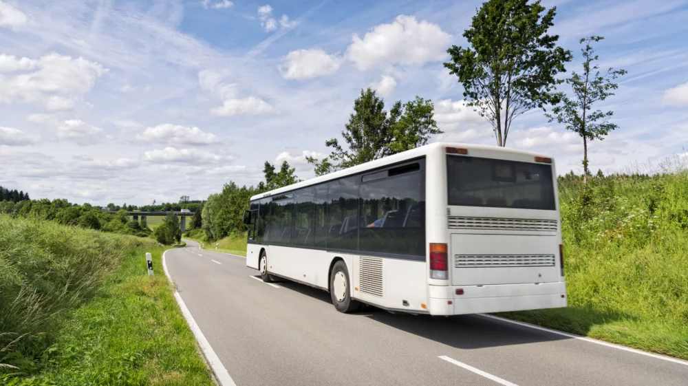 A big white commuter bus driving on a countryside road in the middle of greenery.