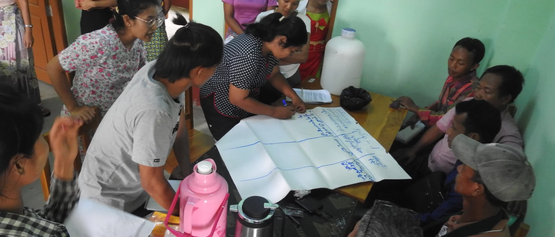 A group of people engaged in a discussion around a table during a wetland conservation training. Participants, including men and women of various ages, are gathered closely, with one person writing on a large sheet of paper placed in the centre.