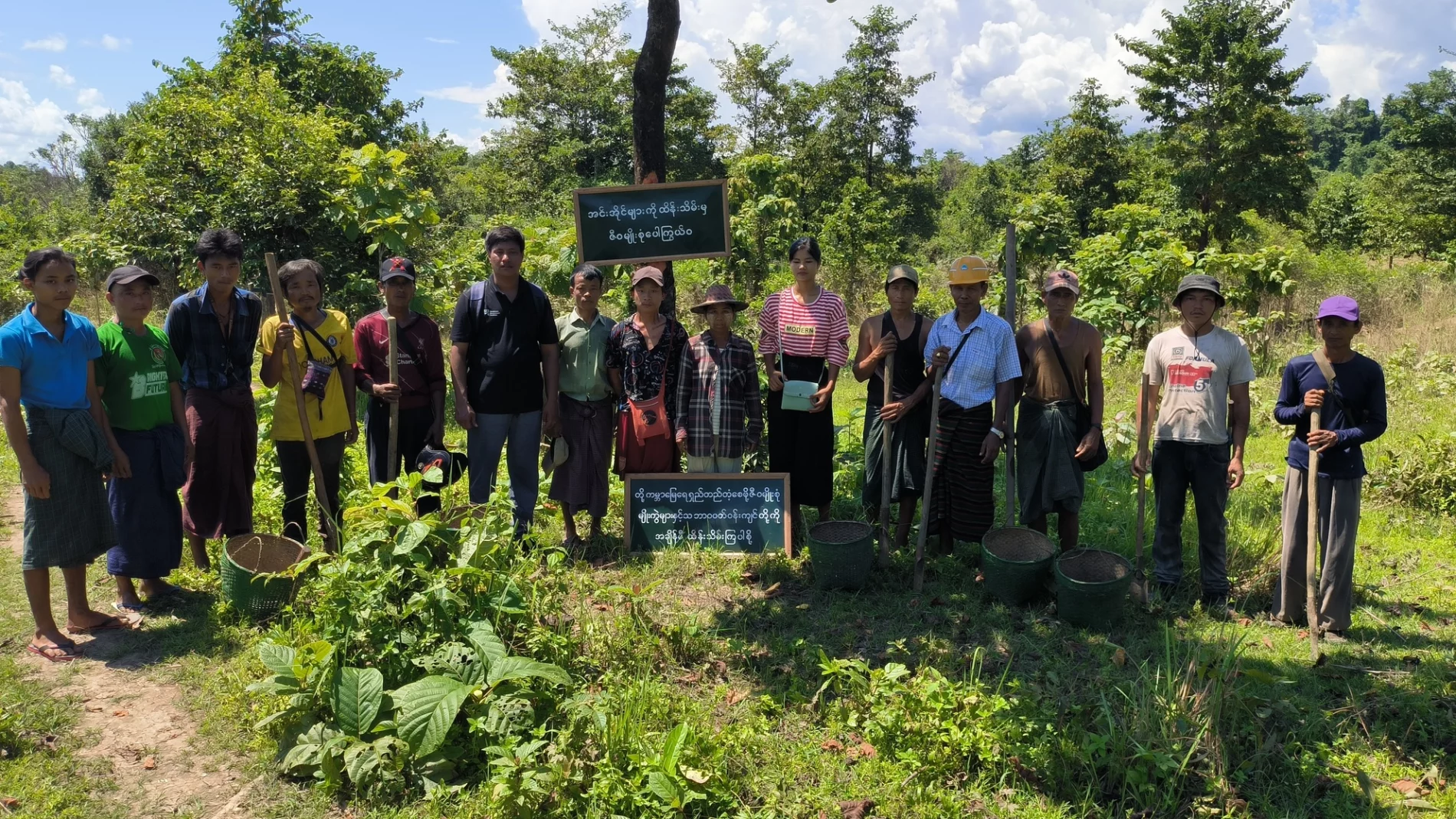 A group of people standing together for a photo during an awareness event and replantation day in Nar Myitter village. They are outdoors in a green, forested area with trees in the background and two signs with local writing displayed prominently.