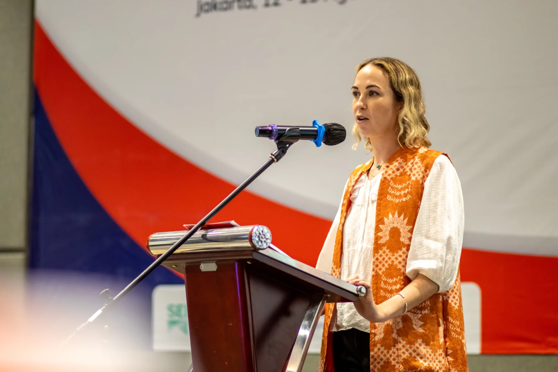 A woman in an orange and white outfit speaks at a podium during the Road Transport Decarbonisation Workshop. A large backdrop with logos is visible behind her.