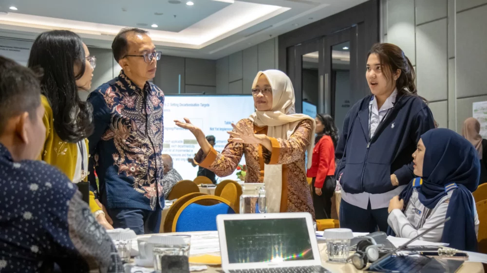 A group of participants stands around a table, engaged in a lively discussion. A woman wearing a headscarf speaks and gestures while others listen intently. Laptops and documents are visible on the table.