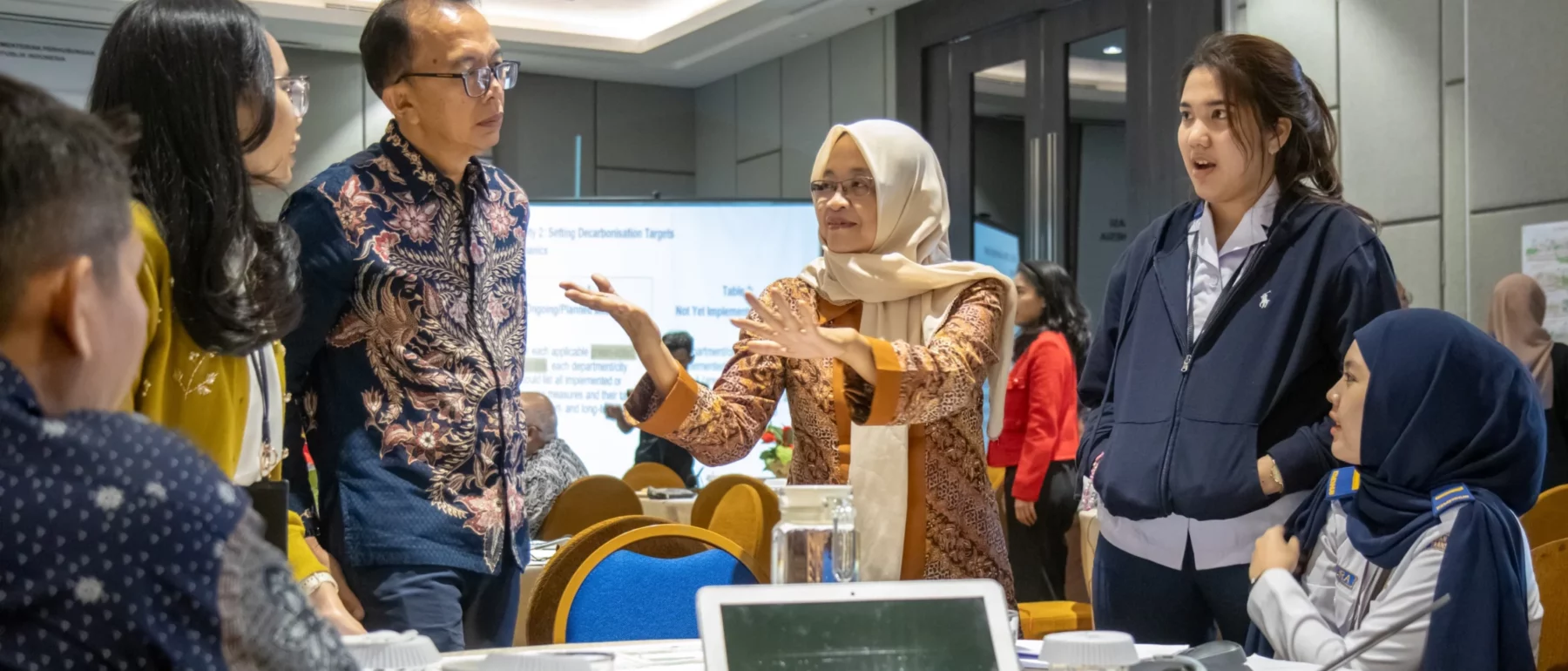 A group of participants stands around a table, engaged in a lively discussion. A woman wearing a headscarf speaks and gestures while others listen intently. Laptops and documents are visible on the table.