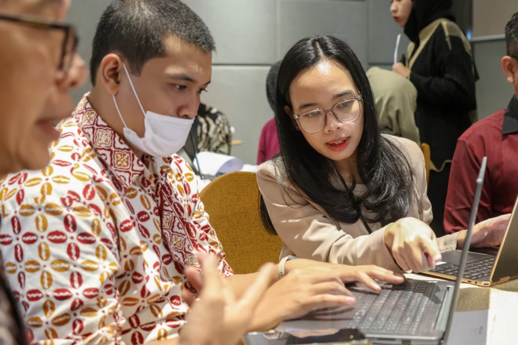 Two workshop participants, one wearing a mask, work together on a laptop. A woman points to the screen while speaking to man sitting next to her.