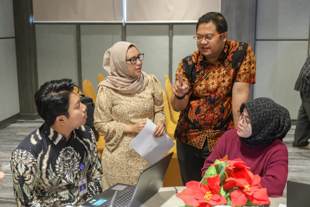 Four participants engage in discussion during a breakout session. One person is gesturing while speaking to others seated around a laptop.