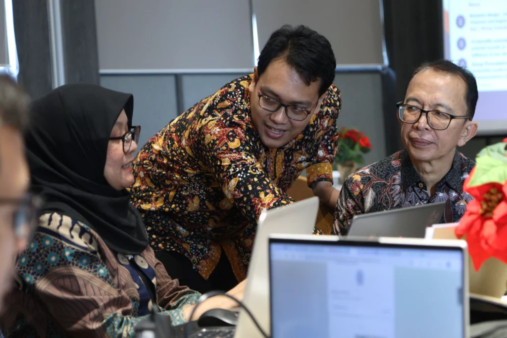 A man leans forward to assist two colleagues sitting at a table with their laptops. All three participants are engaged in a discussion.