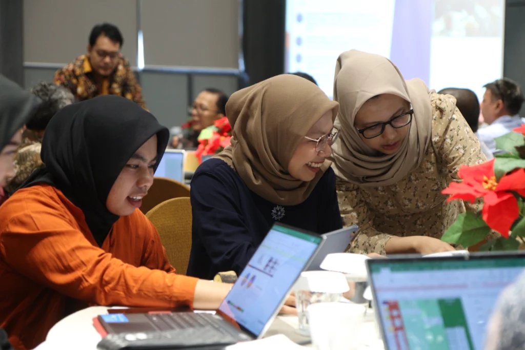 Three women, all wearing headscarves, are gathered around laptops, smiling and discussing.