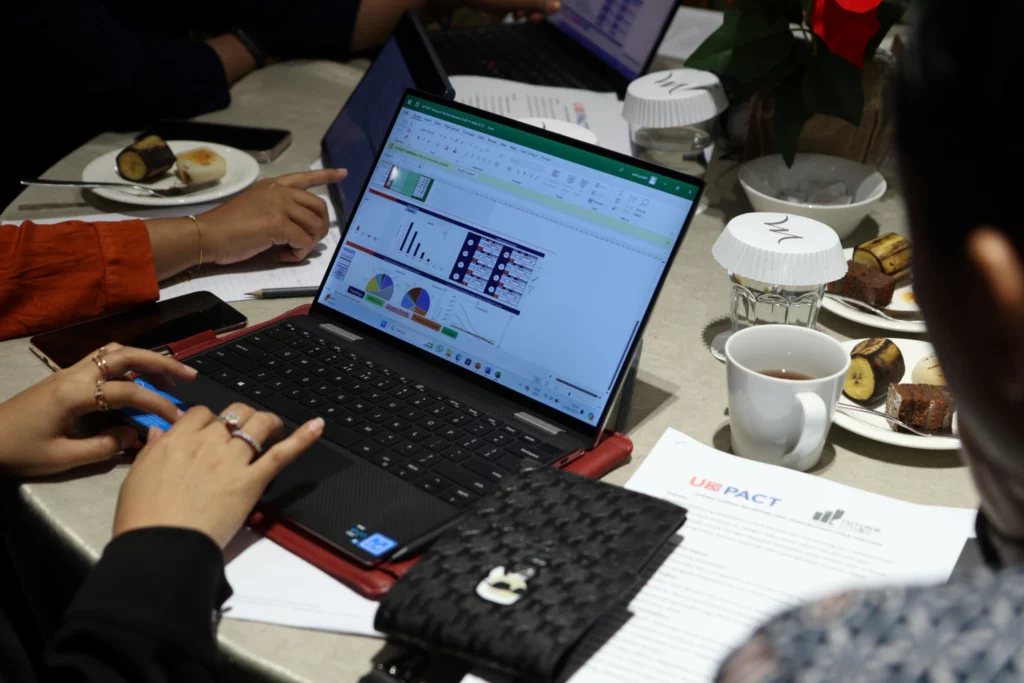 A close-up of a laptop screen displaying charts and graphs from the Road Transport Decarbonisation Pathway (RTDP) tool during a workshop session. Participants' hands are seen working on the laptop, with cups of coffee and snacks on the table.