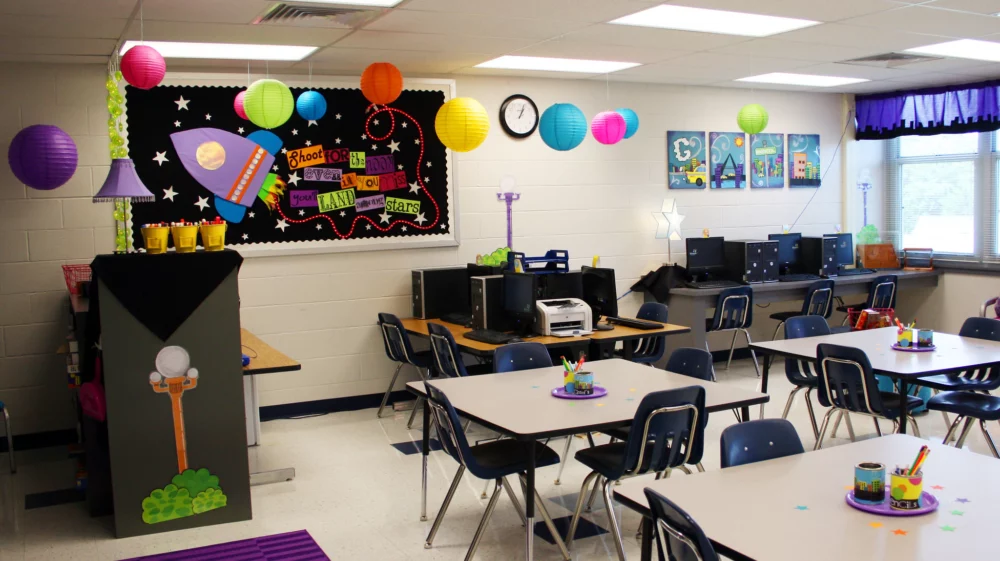 Photo of a tidy classroom with square desks surround by chairs, brightly coloured displays on the walls and hanging from the ceiling and windows visible in one wall.