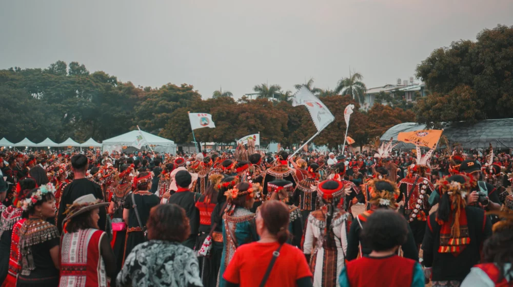 A large group of people in traditional, colourful clothing participating in an event outdoors. The crowd is gathered in a circle, some holding flags, with trees and tents visible in the background under a hazy sky.