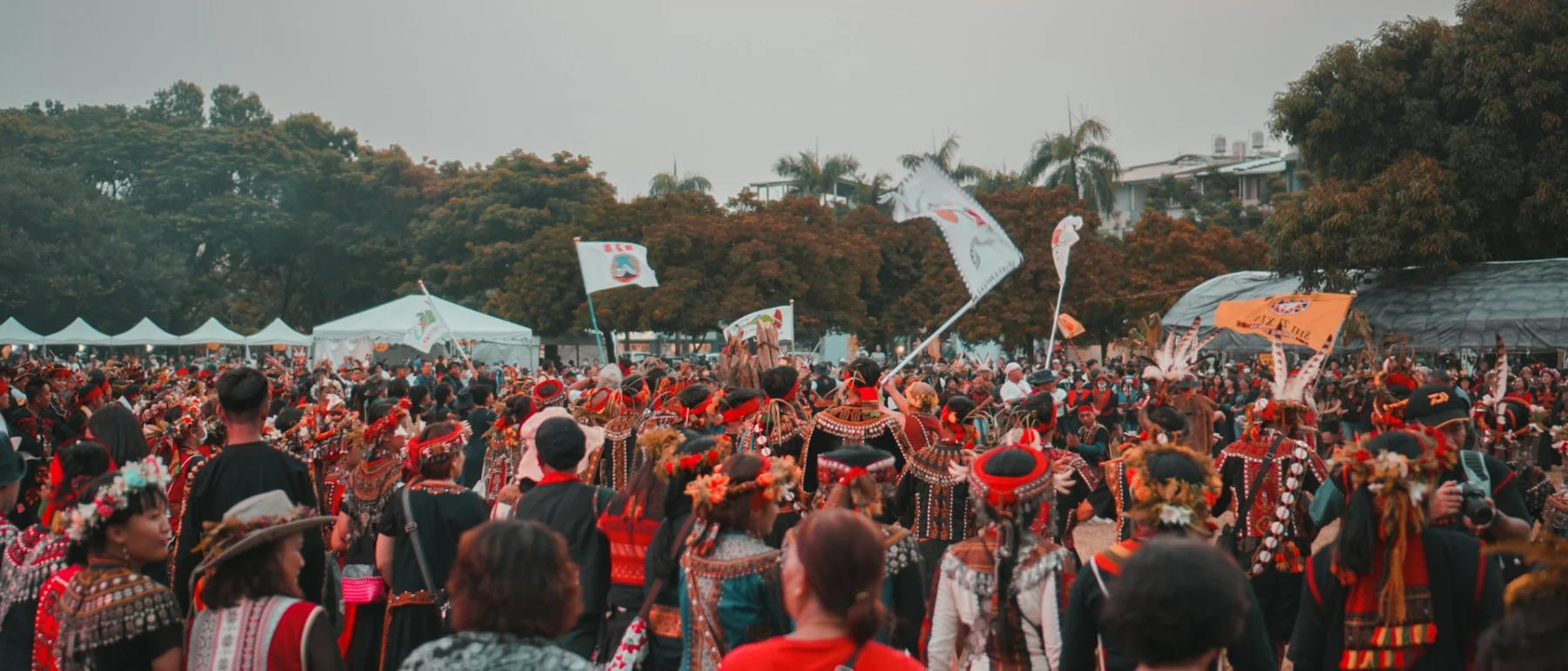 A large group of people in traditional, colourful clothing participating in an event outdoors. The crowd is gathered in a circle, some holding flags, with trees and tents visible in the background under a hazy sky.
