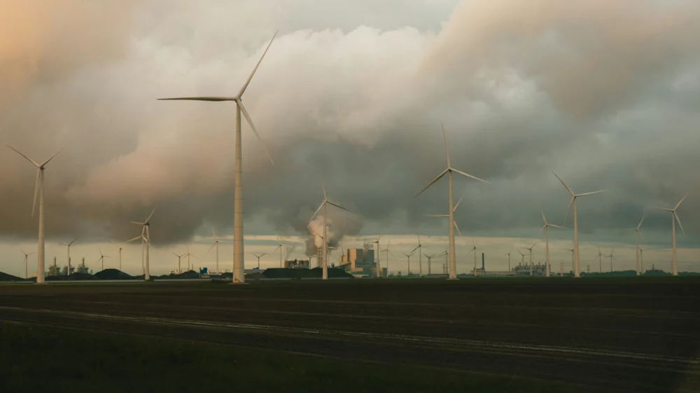 a group of windmills on a cloudy day. In the heart of Eemshaven's industrial harbor, a field of wind turbine stand tall, asserting its presence beneath the stormy skies of the Netherlands. As the sun dips below the horizon, casting an orange glow, a coal-burning power station looms in the background, with thick smoke billowing into the air.