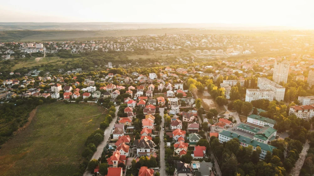 Aerial view of a suburban area with rows of houses featuring red roofs and green spaces, surrounded by trees and greenery. The landscape extends into hills in the background, with the sun casting a warm glow over the scene.
