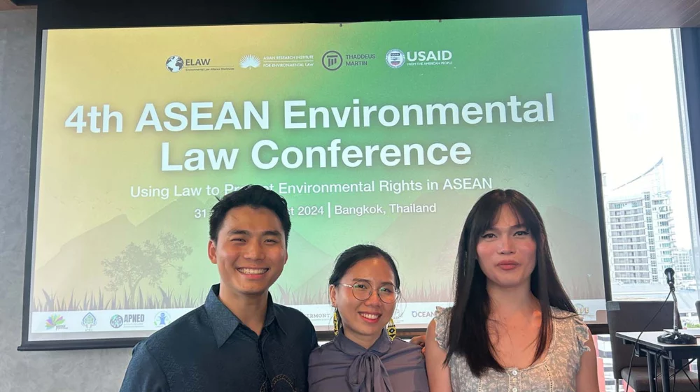 Three youth advocates standing in front of a conference screen displaying “4th ASEAN Environmental Law Conference: Using Law to Protect Environmental Rights in ASEAN.” They are in Bangkok, Thailand, participating in discussions about environmental rights and policies in Southeast Asia.