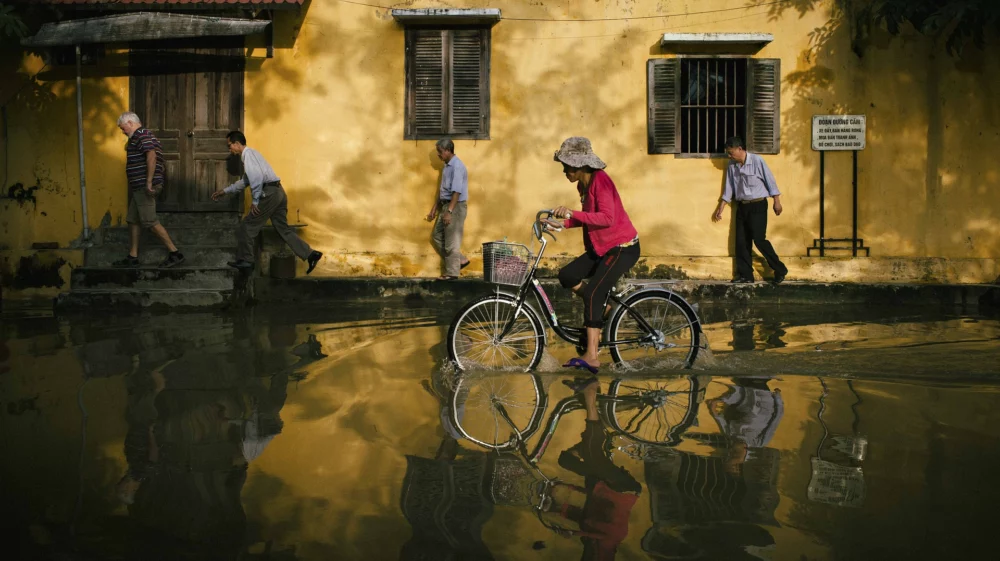 A woman rides a bicycle through a flooded street in Hoi An, Vietnam, while several pedestrians walk carefully along the road. The scene shows reflections in the water from a yellow building.