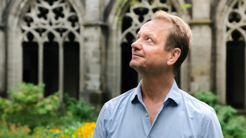 Frank Biermann, in a light blue shirt, looks upward while standing in a garden with stone gothic arches and greenery in the background.