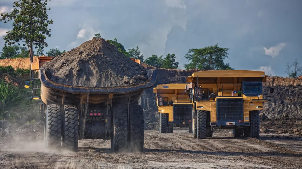 Four yellow trucks drive on a dirt road, with one truck driving away from the viewer and loaded with a pile of coal, and two driving toward the viewer.
