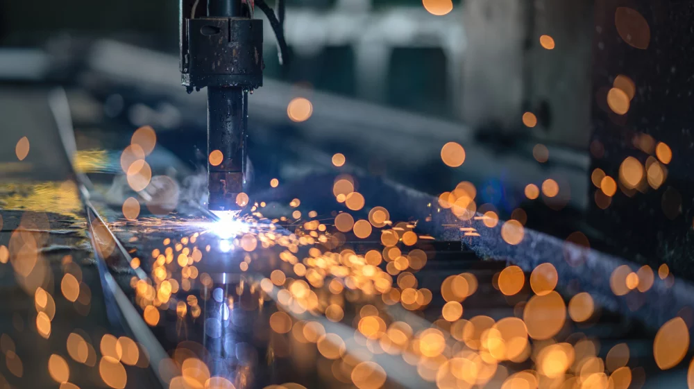 Close-up of a metal cutting process using a high-temperature welding or plasma cutting machine, with bright orange sparks flying in various directions against a blurred background, creating a dynamic and industrial scene.