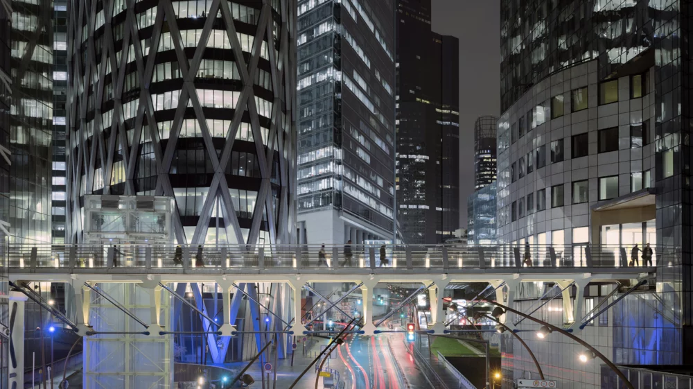 Lit-up black and gray office buildings rise against a dark sky, above a pedestrian bridge, above streaks of lights in blue and red over a street