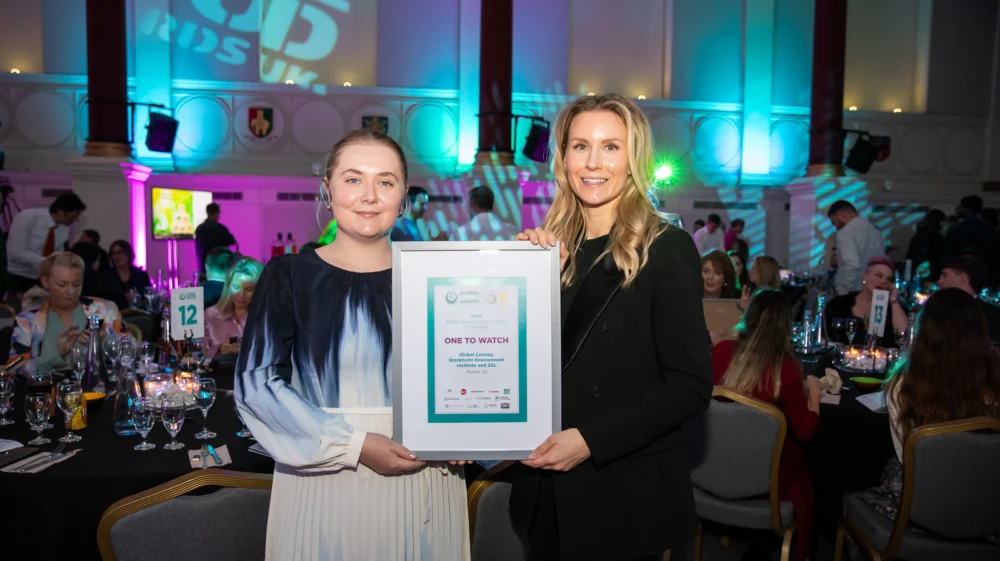 Two women standing in a formal event hall with colourful lighting. The woman on the left is holding a framed award certificate titled “One to Watch,” presented by the Global Good Awards. Seated guests and tables are visible in the background, with ambient purple and green lighting casting a festive atmosphere.