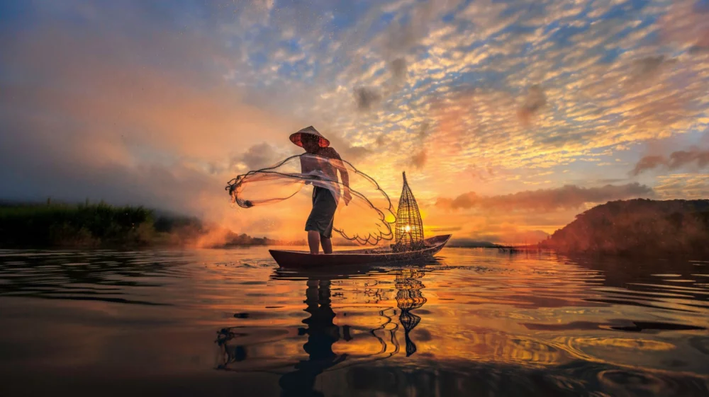 A fisherman standing on a small wooden boat, casting a net into a calm lake or river during a vibrant sunset.