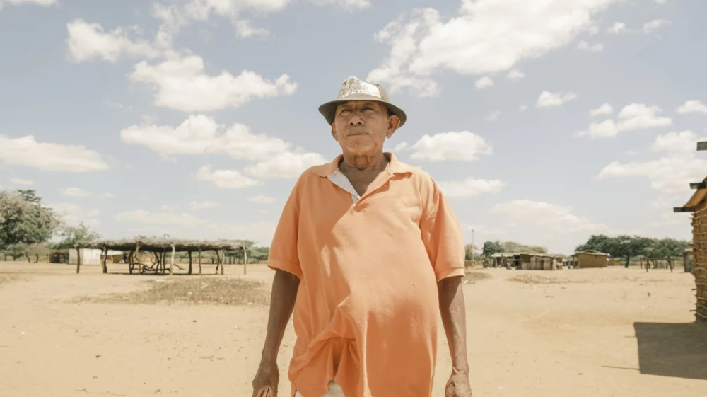 An older man wearing an orange shirt and a hat stands outside in a dry, rural landscape with clear skies.